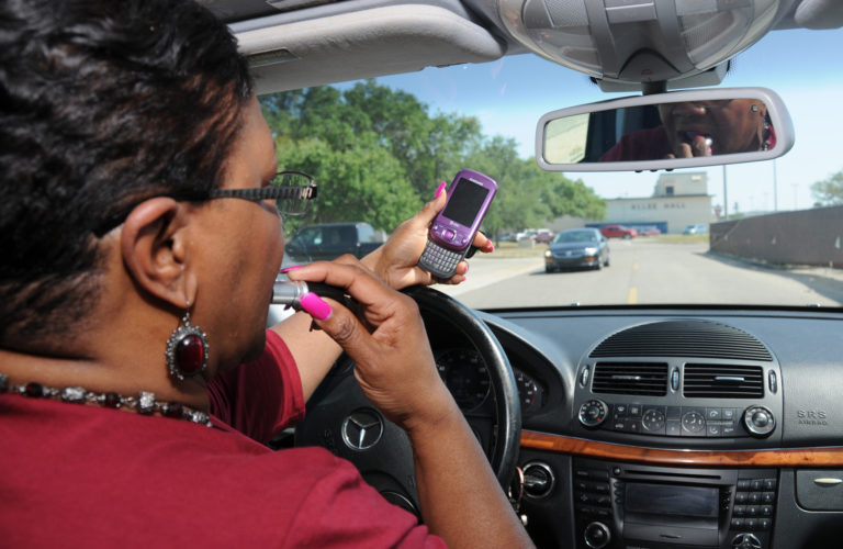 A woman engaging in distracted driving looking at her phone and putting on lipstick