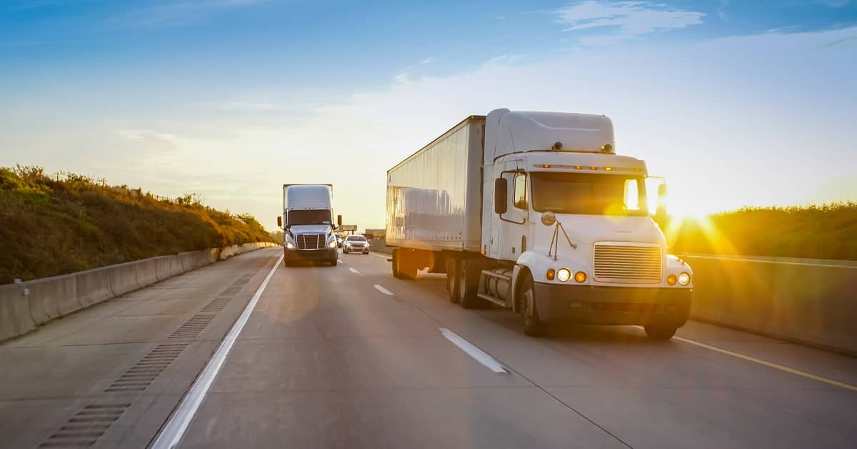 Two white 18 wheeler trucks driving on highway during sunset