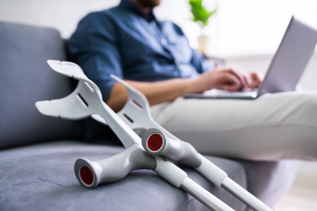 Man with a broken leg sitting on couch with laptop and crutches