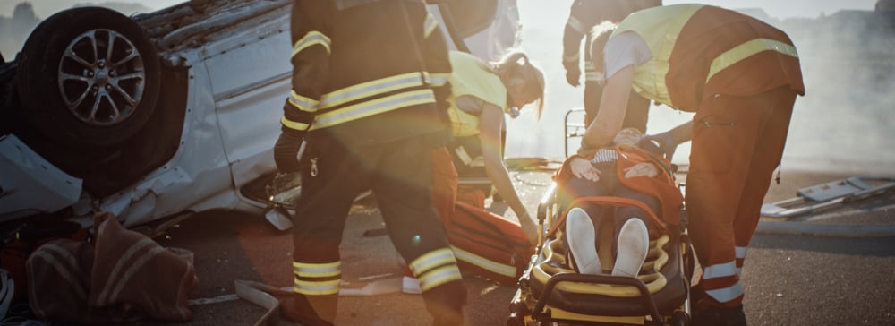 Firefighters and EMTs loading an injured driver onto a stretcher next to a flipped-over car