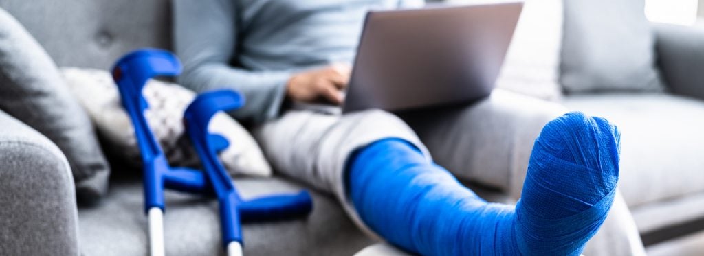 A person with a blue cast and blue crutches sitting on the couch using a laptop
