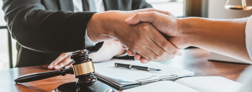Two people shaking hands at a desk inside of a lawyer's office.