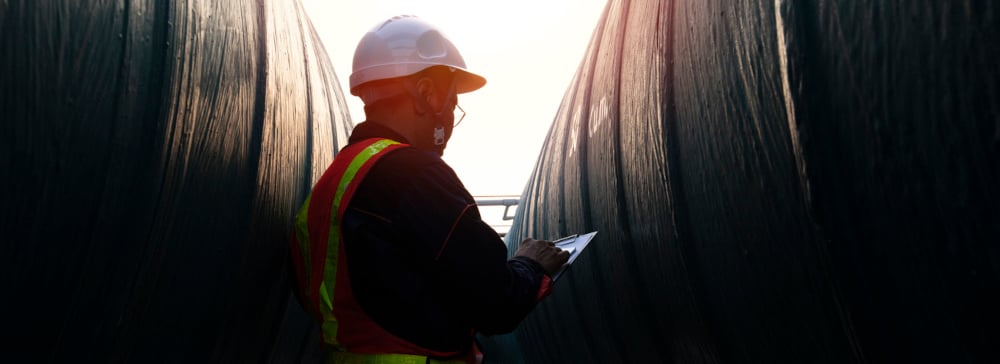 An engineer in a hardhat looking at a clip board between two large pipes