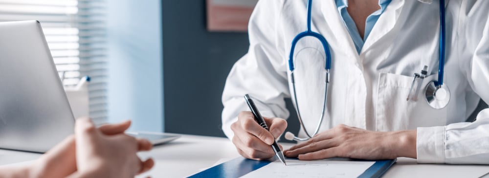 A doctor at a desk with a patient writing a prescription