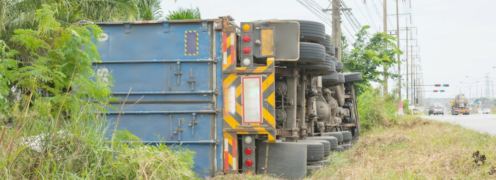 An overturned semi truck in the weeds on the side of the road.