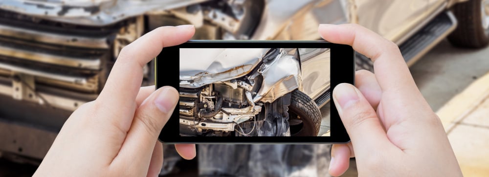A person holding up a smartphone taking a picture of damage to a grey car