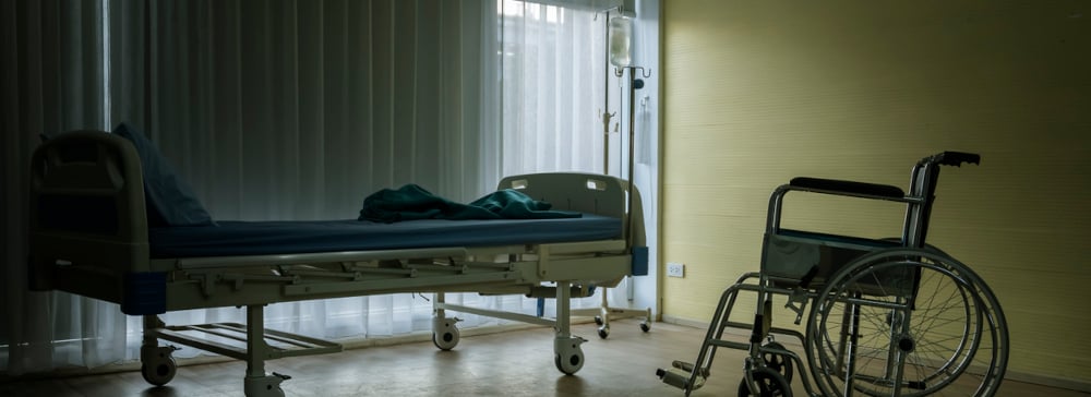 A dark, empty nursing home room with a bed and wheelchair