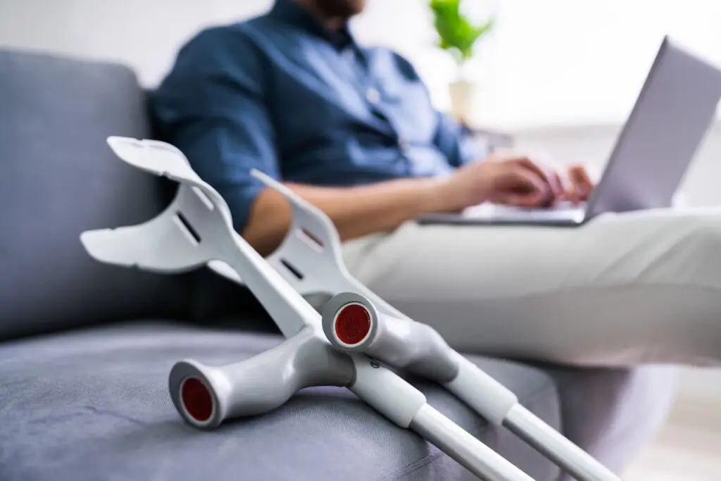 Man with a broken leg sitting on couch with laptop and crutches